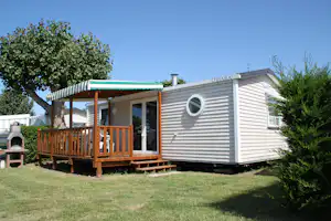 Mobilheim mit Holzterrasse, Markise und Eingangstür, im Hintergrund Bäume und ein Grill am Campingplatz.