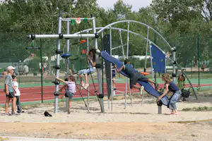 Blaues Klettergerüst mit Rutsche auf einem Spielplatz, Kinder spielen dahinter vor einem grünen Zaun und Bäumen.