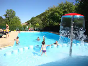 Camping-Schwimmbad mit Wasserfontäne im Vordergrund, Kinder im Becken und Liegen am Rand, Bäume und Hügel im Hintergrund.