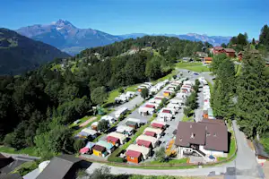 Luftaufnahme eines Campingplatzes im Tal mit Wohnwagen und kleinen Häusern, umgeben von Bäumen und Bergen im Hintergrund.