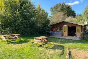Holzunterstand mit Sitzbank und Tisch auf einer grünen Wiese, umgeben von Bäumen und Hecken.