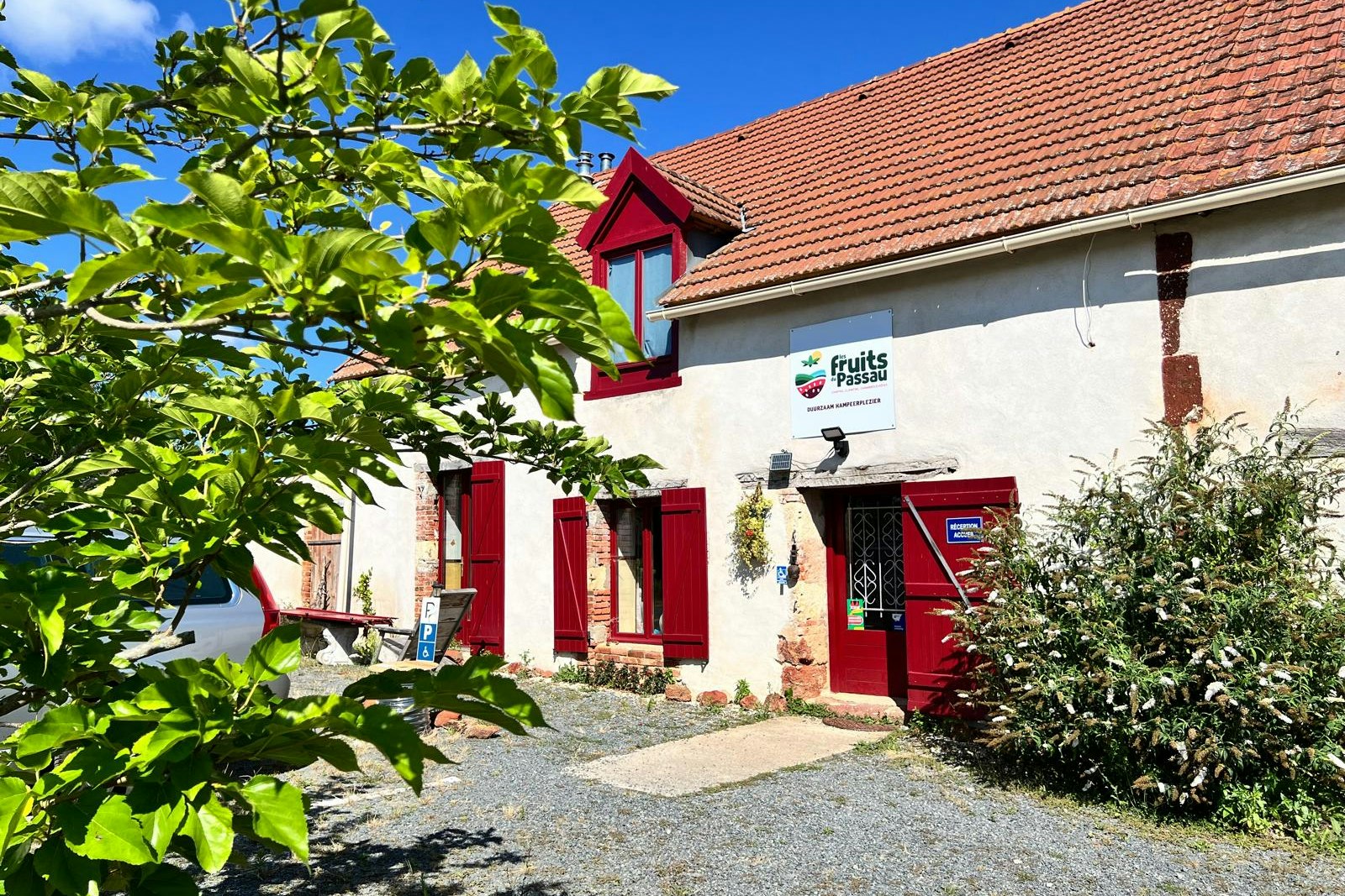 Campinghaus mit rotem Eingang, Schild an der Wand und Kiesweg, umgeben von Büschen und einem Baum im Vordergrund.