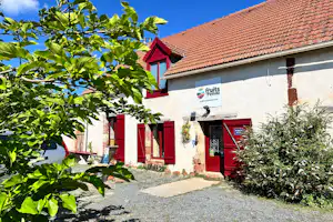 Campinghaus mit rotem Eingang, Schild an der Wand und Kiesweg, umgeben von Büschen und einem Baum im Vordergrund.