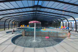 Beheiztes Schwimmbecken unter Glasdach mit Liegen, gelben Schwimmreifen und Kindern im Wasser.