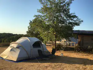 Zelt auf einem Campingplatz mit Campingstühlen und Tisch, daneben eine kleine Holzunterkunft und Bäume im Hintergrund.