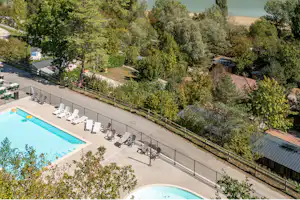 Blick auf den Campingplatz mit blauem Pool, Liegen und Terrasse, umgeben von Bäumen und einem See im Hintergrund.