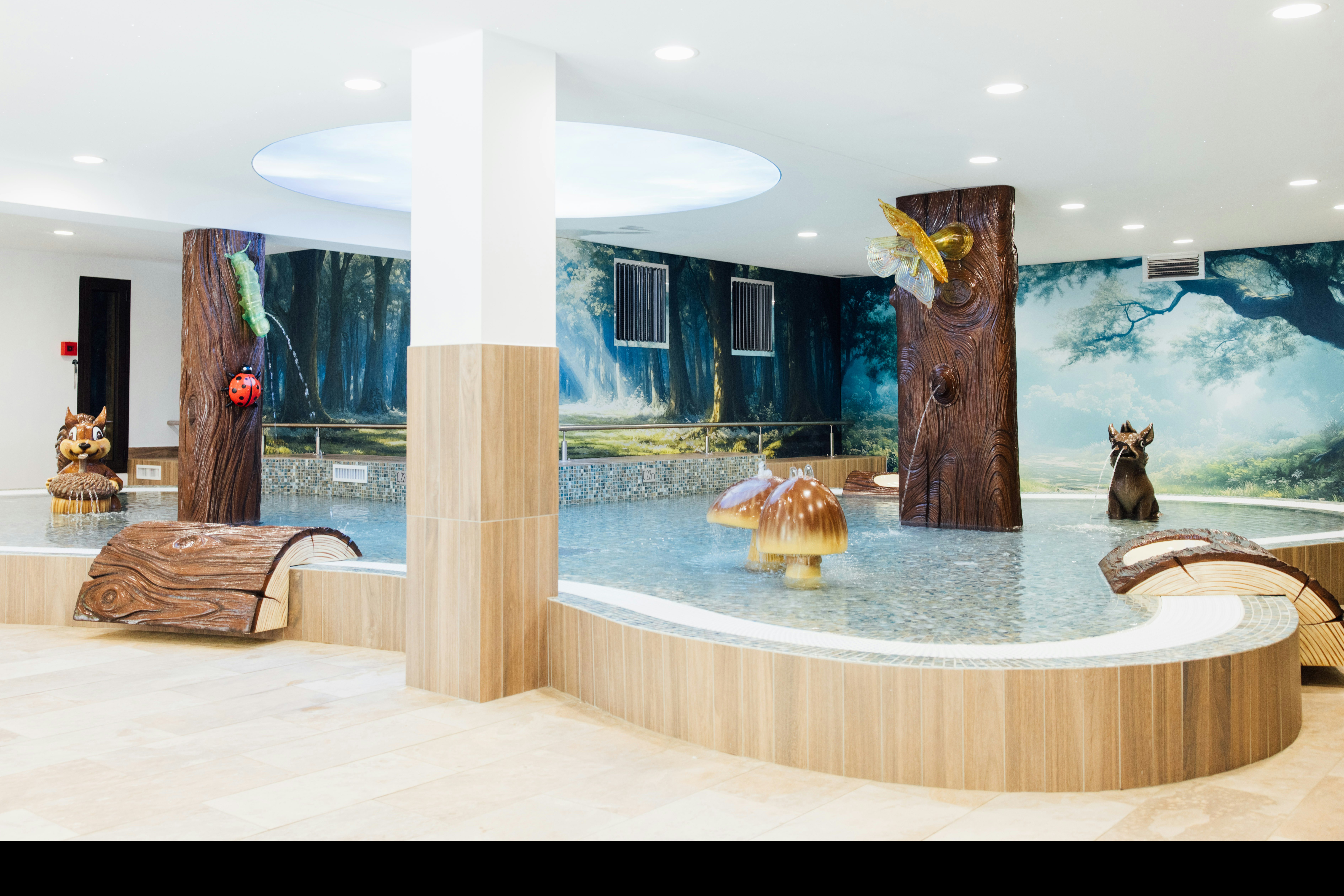 Innenpool mit Holzverkleidung, runder Beckenform und Wasserfontänen, umgeben von Liegen und großen Wandmotiven.