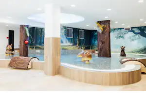 Innenpool mit Holzverkleidung, runder Beckenform und Wasserfontänen, umgeben von Liegen und großen Wandmotiven.