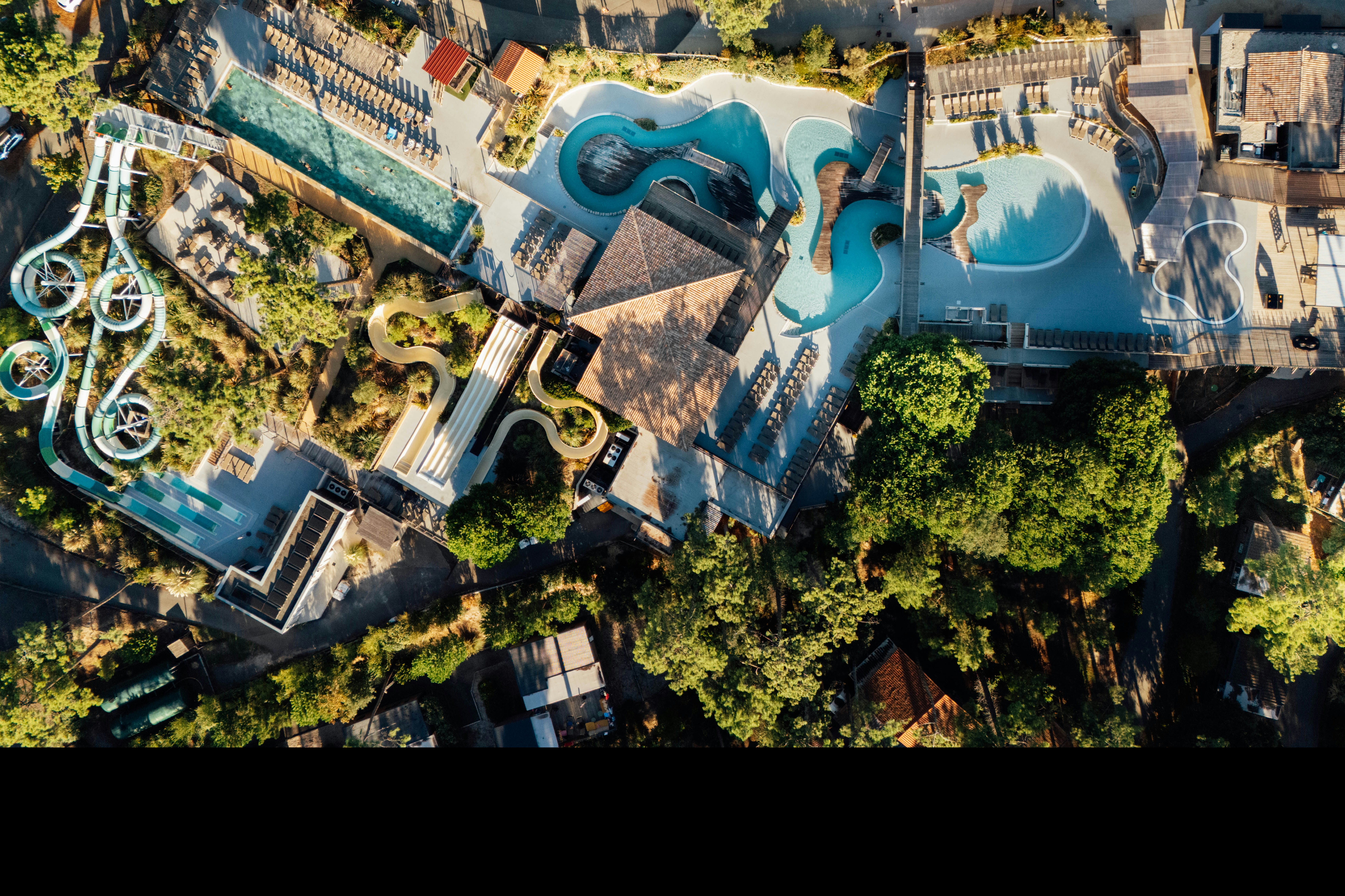 Luftaufnahme eines Campingplatzes mit zwei Pools, Sonnenliegen, Wegen und Gebäuden zwischen Bäumen und Grünflächen.
