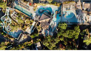 Luftaufnahme eines Campingplatzes mit zwei Pools, Sonnenliegen, Wegen und Gebäuden zwischen Bäumen und Grünflächen.