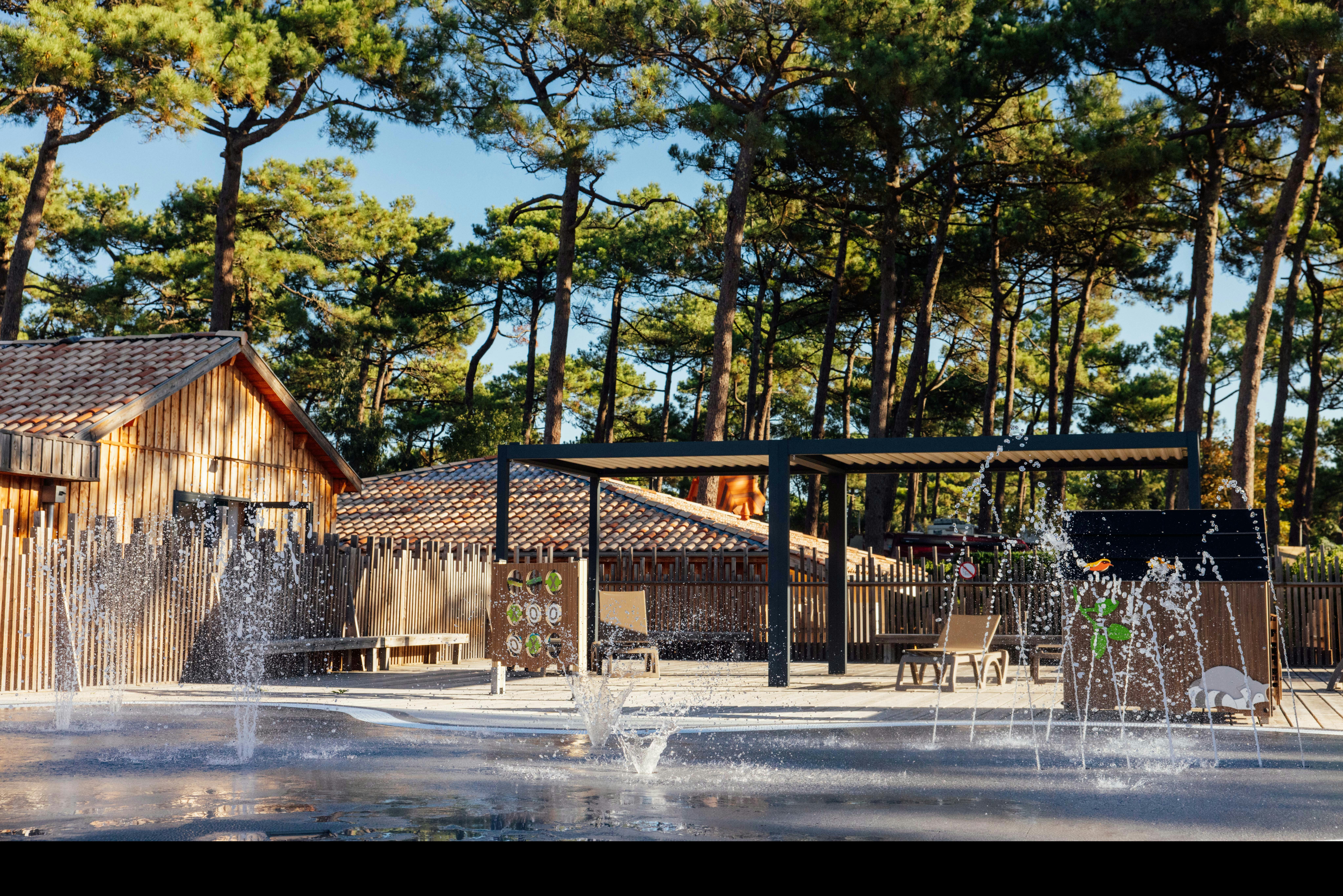 Holzhütte und überdachte Terrasse am Campingplatz, im Vordergrund mehrere Springbrunnen mit Wasserstrahlen vor den Bäumen.