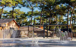 Holzhütte und überdachte Terrasse am Campingplatz, im Vordergrund mehrere Springbrunnen mit Wasserstrahlen vor den Bäumen.