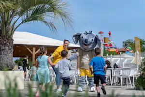 Familie mit Kindern und Maskottchen im Campingbereich, neben weißen Stühlen und einem Zelt mit Palmen im Hintergrund.