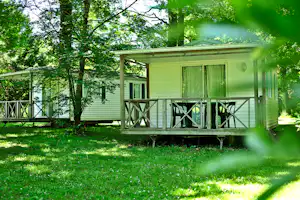 Wohnwagen mit Veranda und Holzgeländer im Grünen, umgeben von Bäumen und Rasen auf einem Campingplatz.