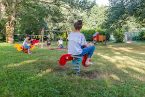 Kinder spielen auf dem Spielplatz mit bunten Schaukeln im Grünen, im Hintergrund Bäume und ein Zaun.