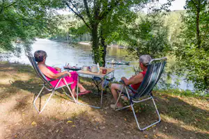 Camping am Fluss: zwei Personen sitzen an einem Tisch mit Kaffeekanne, umgeben von Bäumen und einem Boot im Wasser.