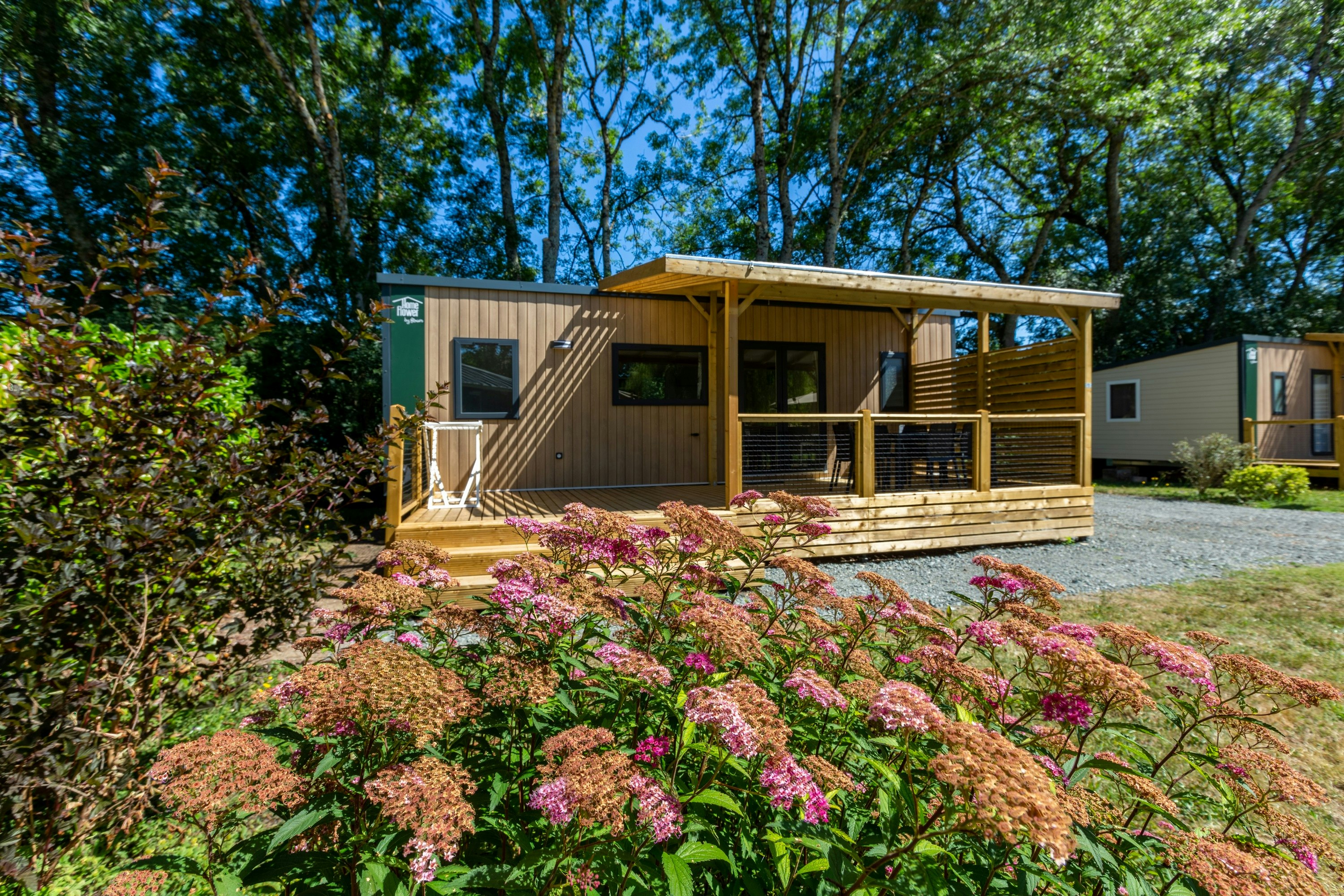 Holzbungalow mit Terrasse, Blumenbeeten und Sitzbereich, umgeben von Bäumen und einem Kiesweg auf dem Campingplatz.