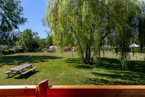 Campingwiese mit großer Weide, Holzbank und Spielbereich im Hintergrund, umgeben von Bäumen und Rasen.