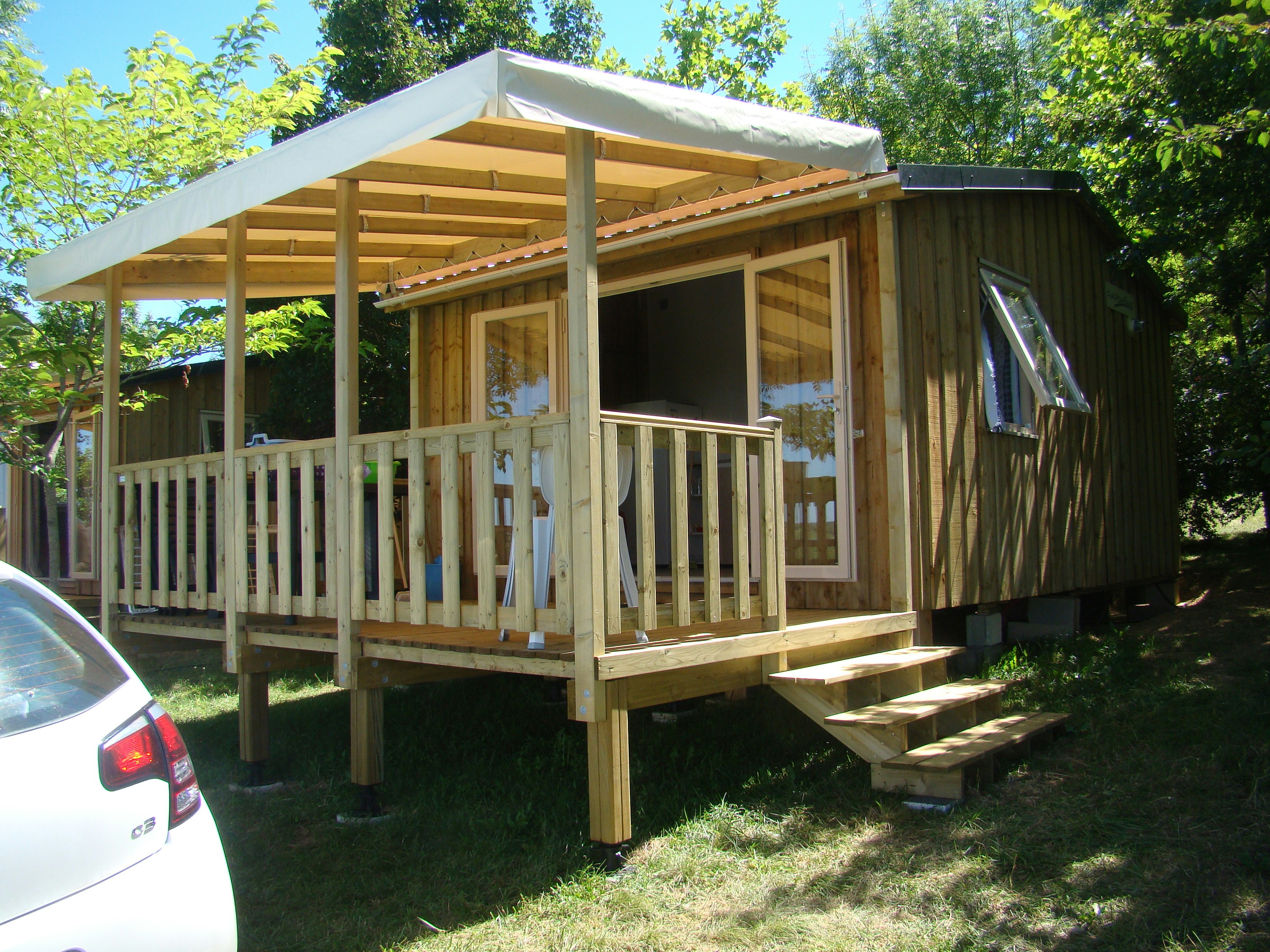 Holz-Mobilheim mit Veranda und Holzgeländer, daneben ein Auto, umgeben von Bäumen und Rasen.