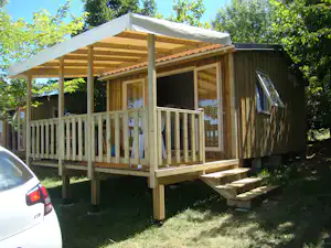 Holz-Mobilheim mit Veranda und Holzgeländer, daneben ein Auto, umgeben von Bäumen und Rasen.