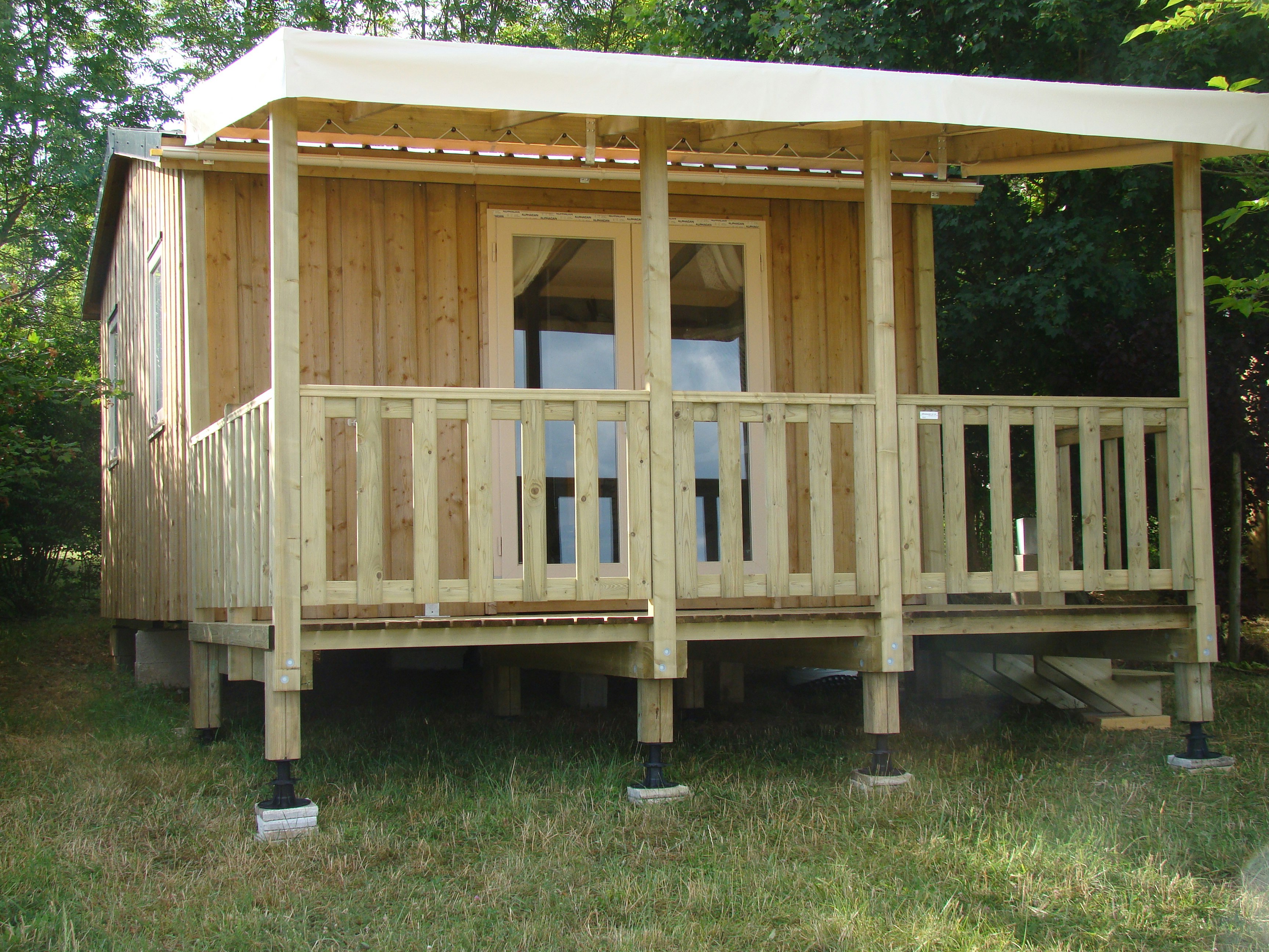 Holzterrasse mit Überdachung und Geländer auf Stelzen, im Grünen mit Bäumen im Hintergrund.