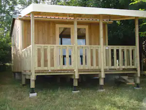 Holzterrasse mit Überdachung und Geländer auf Stelzen, im Grünen mit Bäumen im Hintergrund.