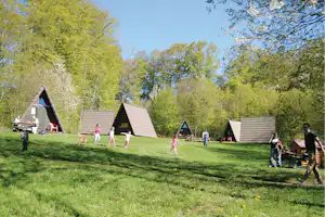 Campingwiese mit mehreren Holzhütten, Spielbereich mit bunten Stühlen und Tischen, umgeben von Bäumen und blauem Himmel.