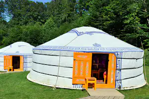 Jurtenzelt mit orangefarbenem Eingang auf einer Holzterrasse, umgeben von dichtem Grün im Campingbereich.