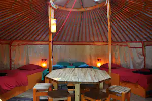 Innenansicht eines Zelts mit Holzbetten, Tisch und Sitzbänken, warmen Lampen und gestreifter Zeltplane.