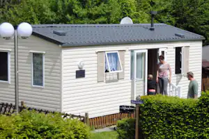 Mobilheim mit Terrasse und großen Fenstern, davor Hecke und Rasen, im Hintergrund grüner Wald.