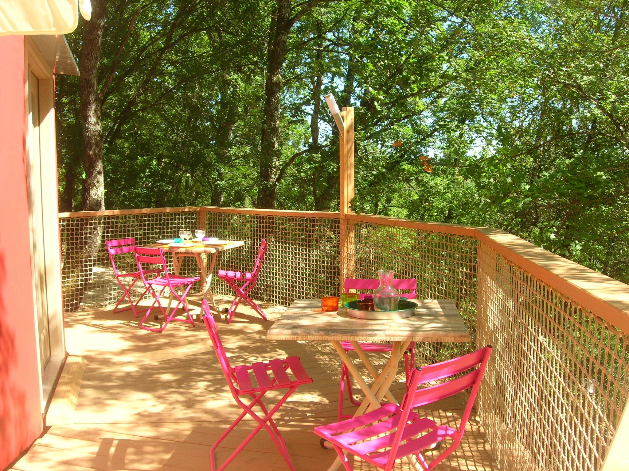 Camping-Terrasse mit bunten Klappstühlen und Tisch, umzäunt mit Holzgeländer, im Hintergrund Bäume im Grünen.