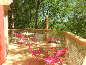 Camping-Terrasse mit bunten Klappstühlen und Tisch, umzäunt mit Holzgeländer, im Hintergrund Bäume im Grünen.