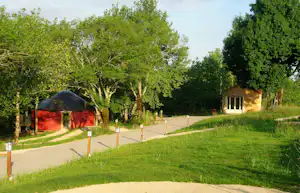 Campingplatzweg mit Rasen und Bäumen, links ein Zelt mit dunkler Plane, rechts ein kleines Gebäude im Grünen.