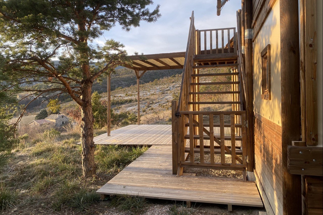 Holzterrasse am Campinghaus mit Treppe, Geländer und Bank, daneben Kiefern und ein Blick auf den Hügel im Hintergrund.