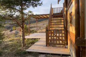 Holzterrasse am Campinghaus mit Treppe, Geländer und Bank, daneben Kiefern und ein Blick auf den Hügel im Hintergrund.