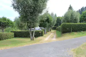 Campingplatz mit Kiesweg, Hecken und Bäumen; im Hintergrund Zelte und Wohnwagen auf Stellplätzen.