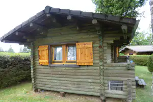 Holzblockhaus mit großen Fenstern, überdachter Terrasse und Bank am Rand eines Campingplatzes, umgeben von Bäumen und Rasen.