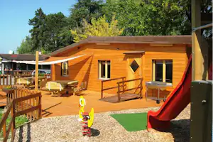 Holzbungalow mit Terrasse, Sitzbank und Spielgerät im Garten, umgeben von Bäumen und Kiesweg.