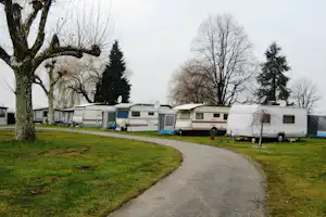 Reihe von Wohnwagen und Caravans auf einem Campingplatz, gepflasterter Weg, Rasen und Bäume entlang der Stellplätze.