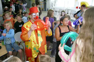 Clown mit roter Nase und Luftballon steht vor Kindern in einem hellen Campinggebäude, Stühle und Tische im Hintergrund.