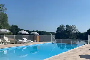 Blauer Camping-Pool mit Liegen am Rand, dahinter Zäune und mehrere weiße Sonnenschirme vor Bäumen.