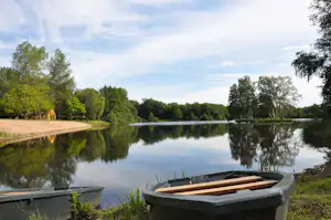 Ruderboot am Ufer eines ruhigen Sees, dahinter Bäume und ein Campingplatz mit Steg und Wegen.