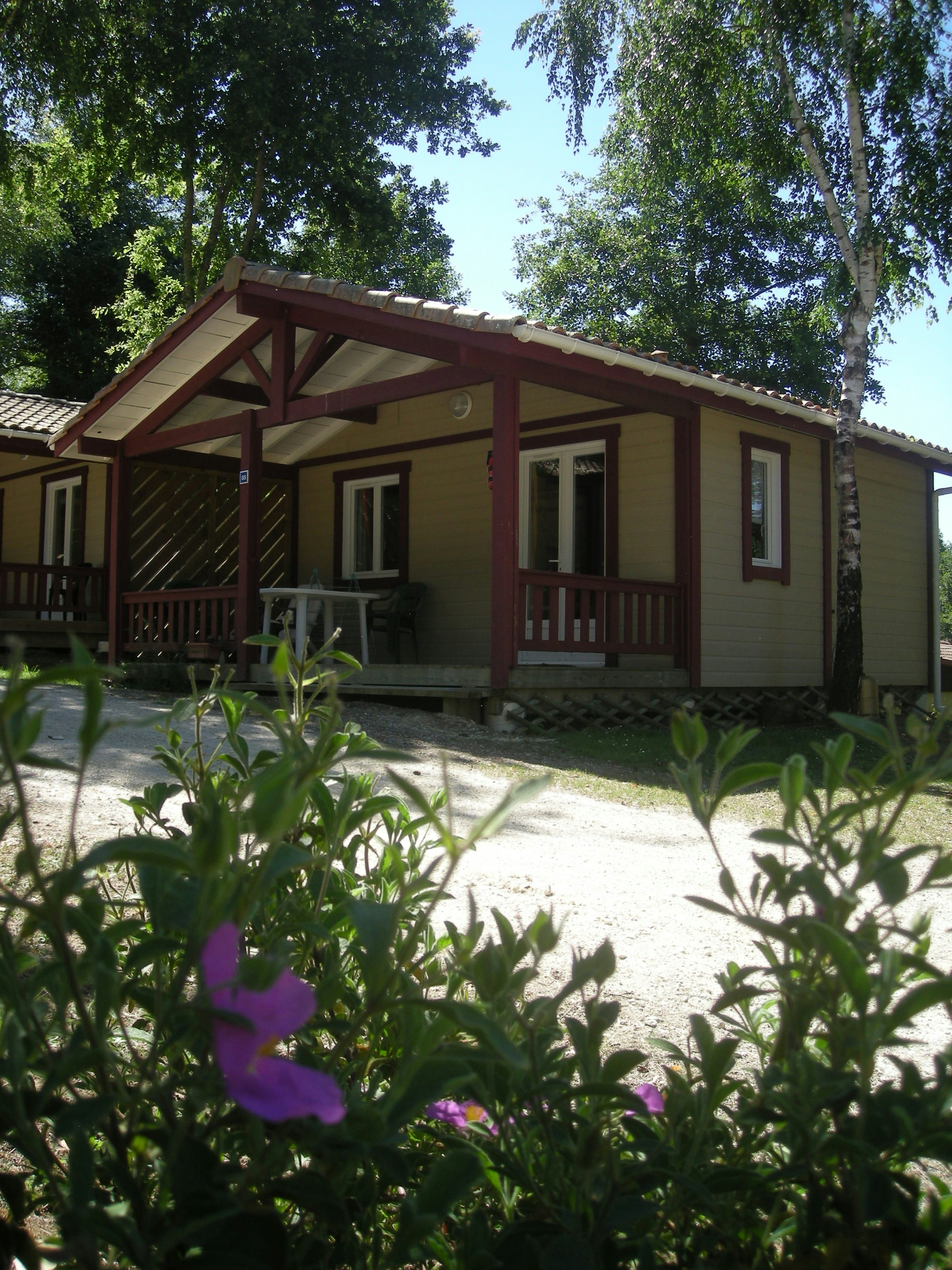 Mobilheim mit Veranda und Holzgeländer, davor Kiesweg und Blumenbeete, umgeben von Bäumen im Campingbereich.