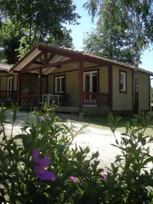 Mobilheim mit Veranda und Holzgeländer, davor Kiesweg und Blumenbeete, umgeben von Bäumen im Campingbereich.