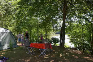 Zeltplatz am Fluss: Zelt, Campingstühle und Tisch unter Bäumen, im Hintergrund Wasser und weitere Zelte.