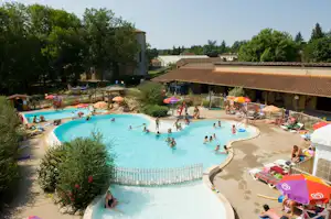 Großes Camping-Schwimmbecken mit mehreren Liegen, Sonnenschirmen und Gebäuden im Hintergrund, umgeben von Bäumen und Wegen.