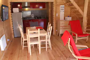Holzhütte mit Küchenzeile, Esstisch und Stühlen, roter Sitzbank sowie Schränken unter dem Dachbalken.