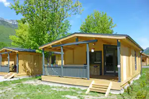 Holzbungalow mit Veranda, Sitzbank und Geländer auf dem Stellplatz, umgeben von Bäumen und grünem Rasen.