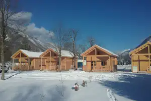Holzhütten mit Veranda und Treppen im Schnee, dahinter Berge und Bäume vor blauem Himmel.