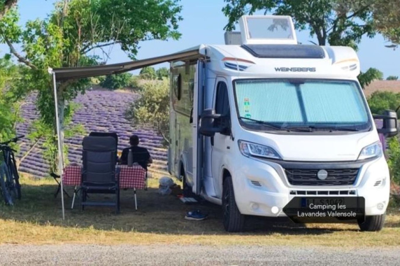 Wohnmobil auf einem Campingplatz mit Markise, davor zwei Campingstühle und Tisch, Bäume und Hecke im Hintergrund.